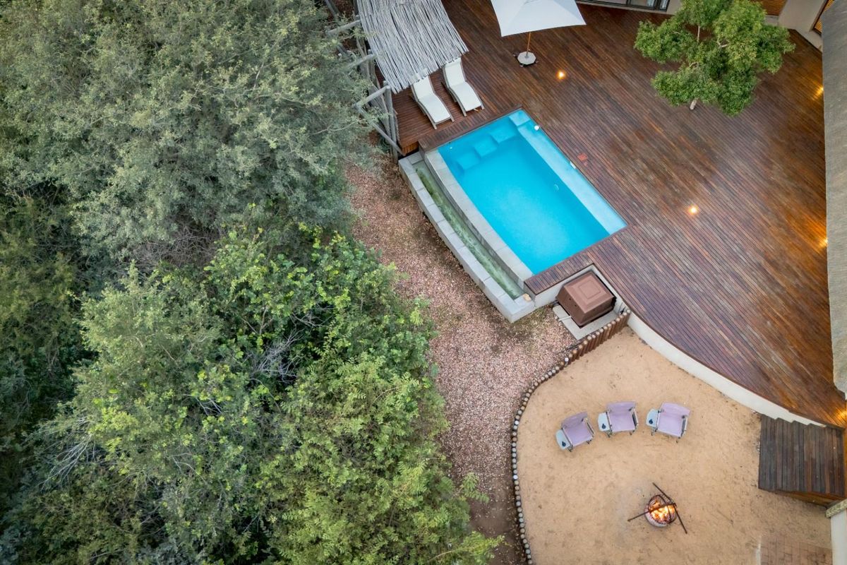 Aerial view of a property featuring a swimming pool, extensive timber decking, and a fire pit area set within a natural landscape.