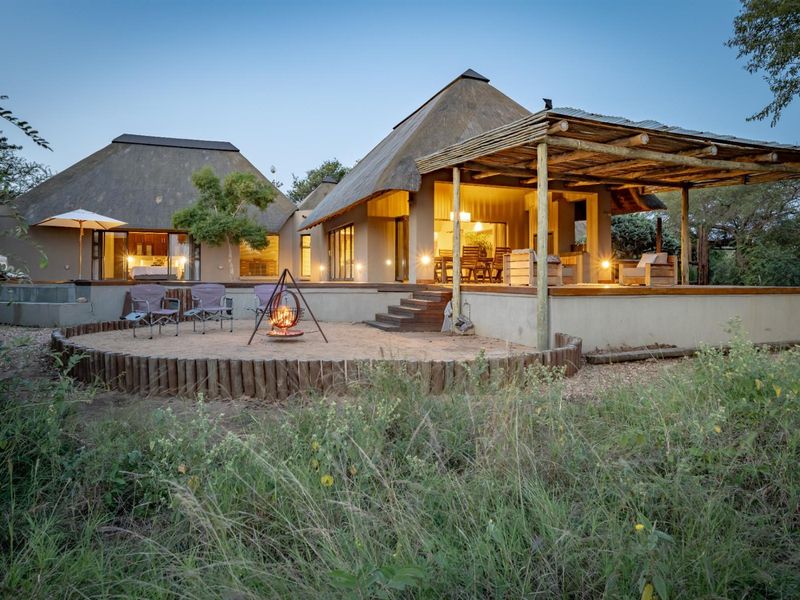 Rear view of a thatched-roof house featuring a raised wooden deck, covered patio, and a sand fire pit area in the garden.