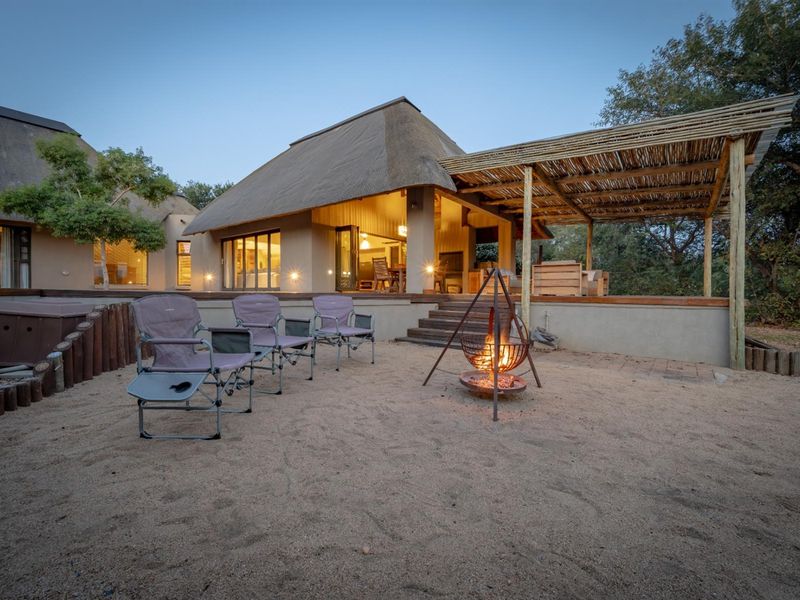 Rear view of a thatched-roof house featuring a raised timber deck, covered pergola, and a sandy outdoor fire pit area.
