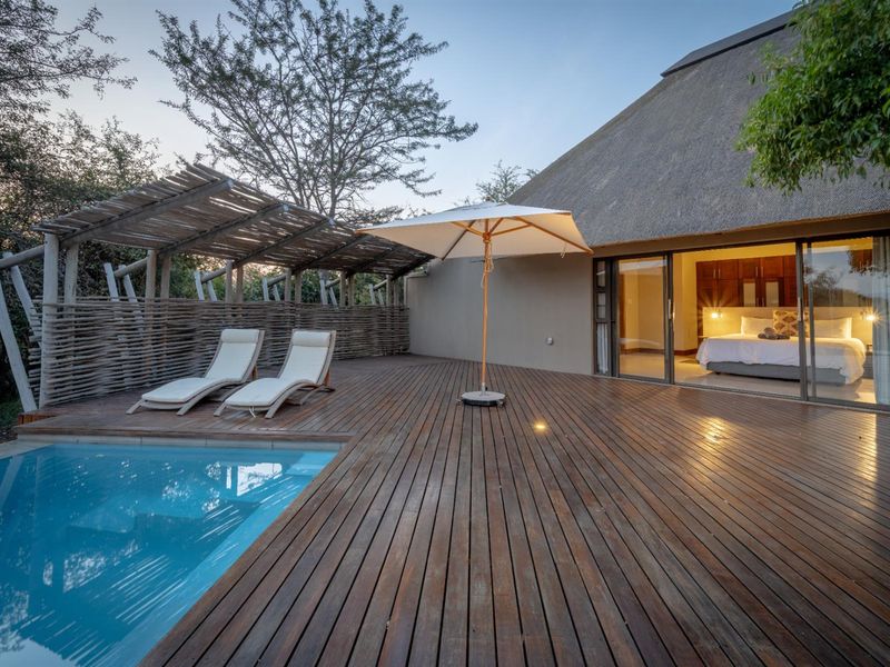 Outdoor pool area featuring extensive timber decking, a thatched roof structure, and seamless access to the interior bedroom through large sliding glass doors.