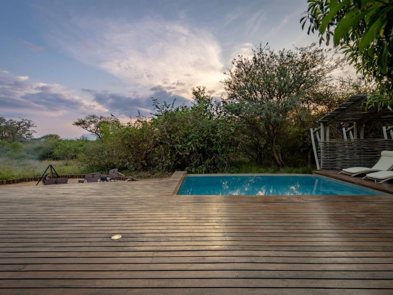 Outdoor pool area featuring extensive timber decking, a swimming pool, and a thatched gazebo set against a natural bushveld backdrop.