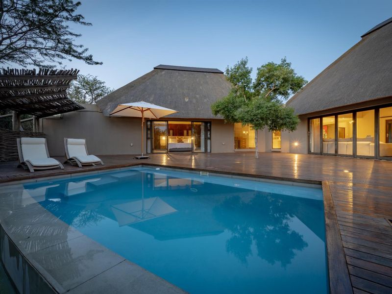 A beautifully maintained outdoor pool area featuring extensive timber decking, thatched roof structures, and seamless indoor-outdoor flow through large sliding glass doors.
