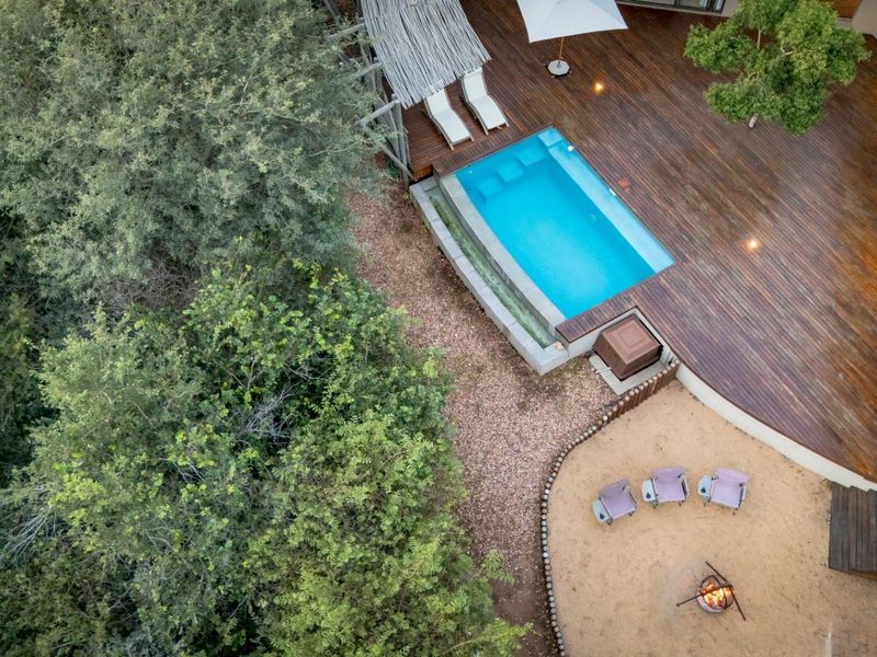 Aerial view of a property featuring a swimming pool, extensive timber decking, and a fire pit area set within a natural landscape.