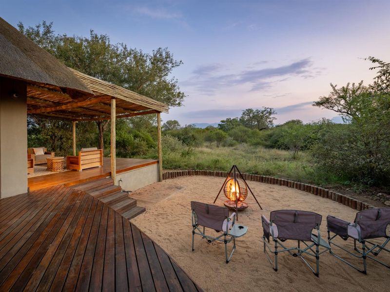 A beautiful outdoor entertainment area featuring a covered thatched patio with timber decking and a dedicated sand fire pit area overlooking the bushveld.