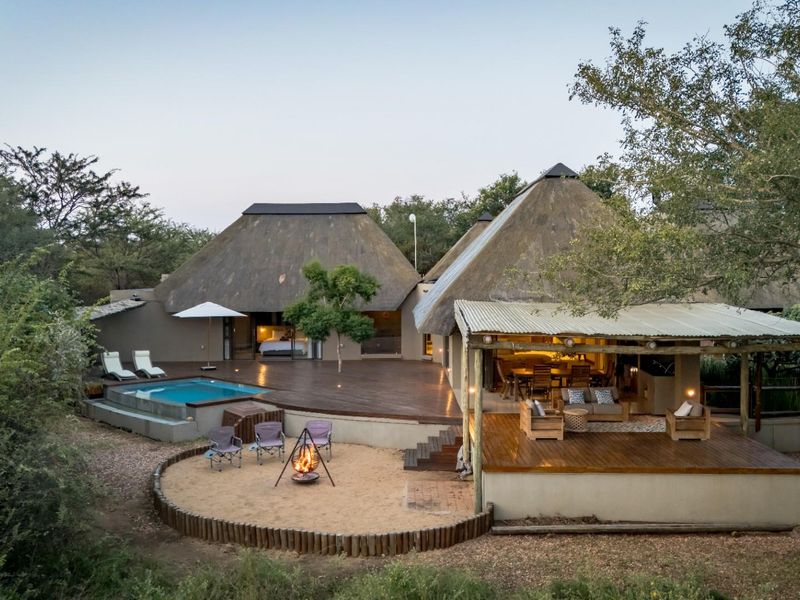 Aerial view of a rear exterior featuring a thatched roof dwelling, timber decking, a swimming pool, and a dedicated fire pit area.