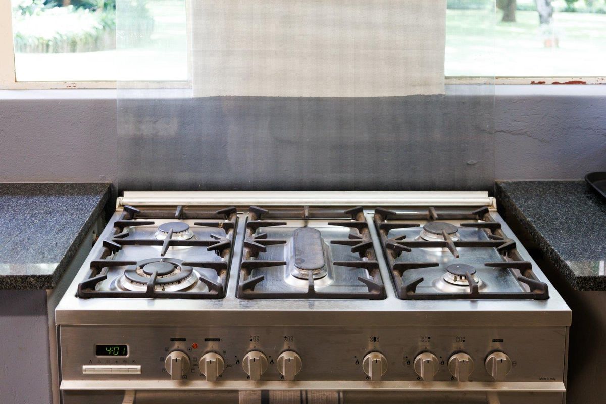 A stainless steel gas hob and oven unit set into dark granite countertops, positioned beneath a window with a glass splashback.