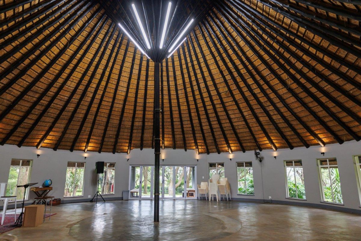 Spacious circular event hall featuring a high vaulted thatched roof, polished concrete flooring, and large windows providing natural light and garden views.