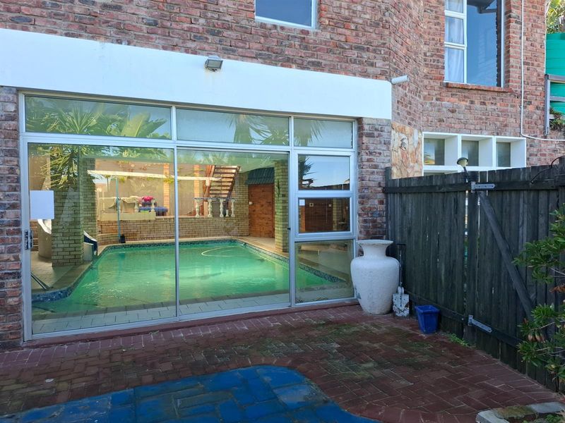 An indoor swimming pool viewed through large sliding glass doors from a paved rear patio area with a timber boundary fence.