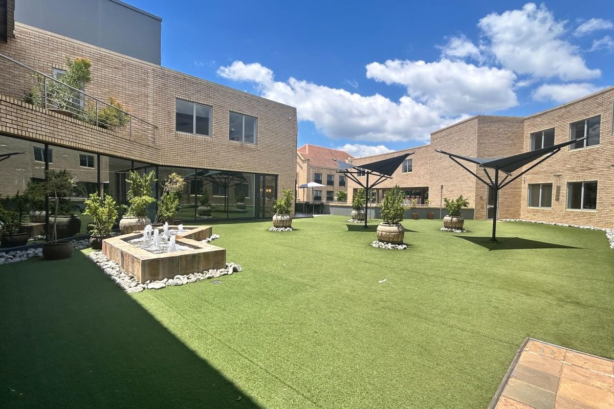 A well-maintained commercial courtyard featuring artificial turf, a central water fountain, and modern brick building facades with integrated shade structures.