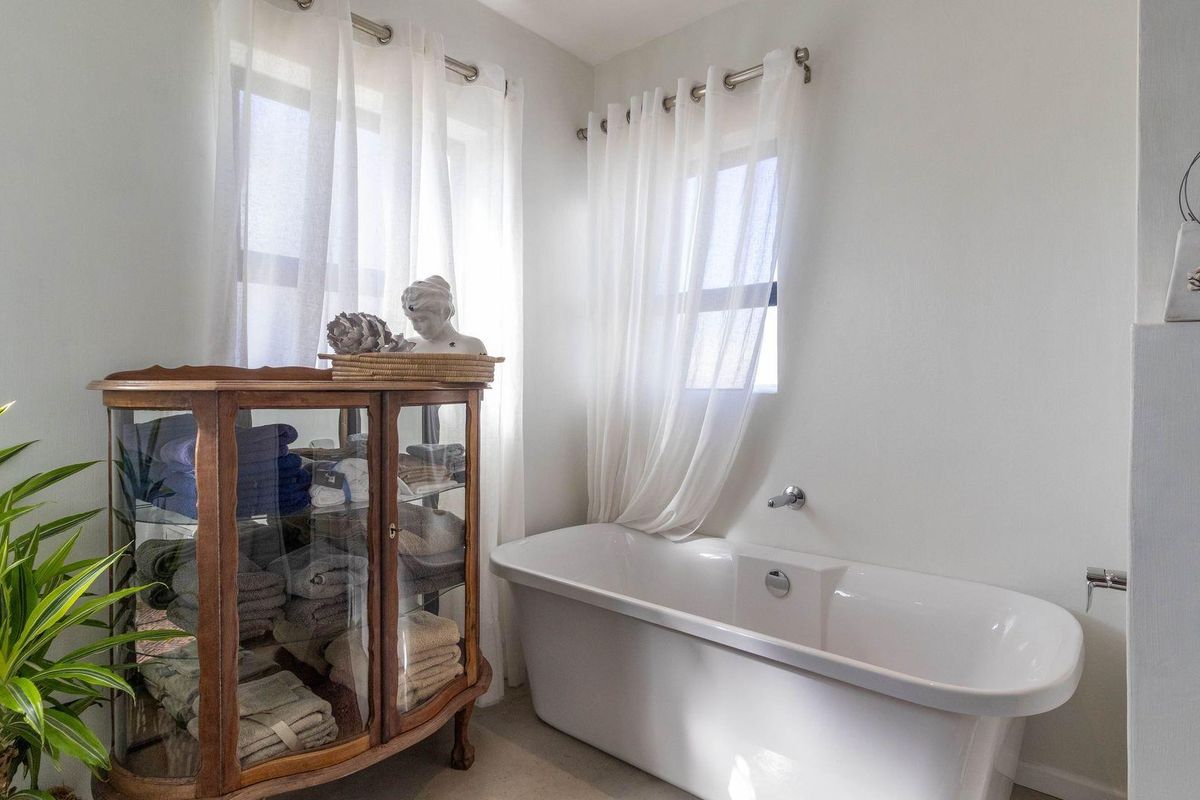 A bright bathroom featuring a modern freestanding bathtub positioned beneath dual windows with sheer curtains.