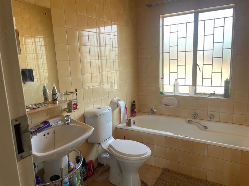 A dated bathroom featuring a pedestal basin, toilet, and built-in bathtub with yellow wall tiling and security bars on the window.
