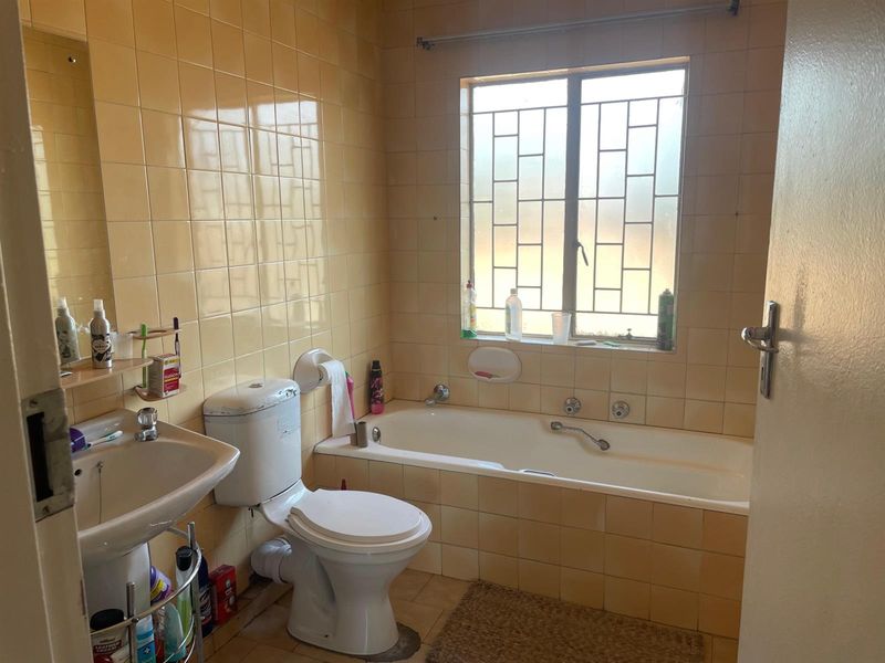 A dated bathroom featuring a pedestal basin, toilet, and built-in bathtub with yellow wall tiling and security bars on the window.