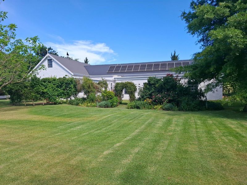 A well-maintained farm residence featuring a white exterior, a pitched roof, and a large array of roof-mounted solar panels set within a lush garden.