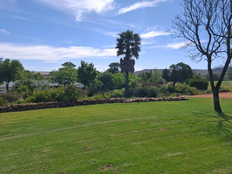 A well-maintained expansive lawn area featuring a stone retaining wall, mature trees, and established garden landscaping on a farm property.