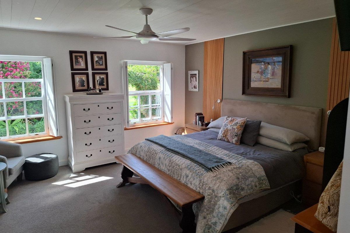 A well-maintained bedroom featuring timber-paneled accent walls, recessed ceiling lighting, a modern ceiling fan, and large windows with internal shutters overlooking the garden.