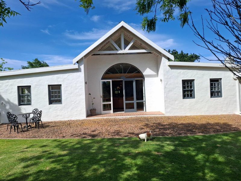 A charming farm-style residence featuring a white stucco facade, arched glass entrance, and a paved patio area overlooking a well-maintained garden.