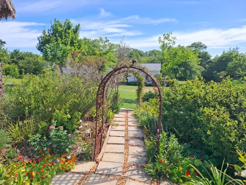 A charming garden pathway featuring a decorative wrought iron archway leading through lush, well-maintained flower beds and greenery on the farm property.