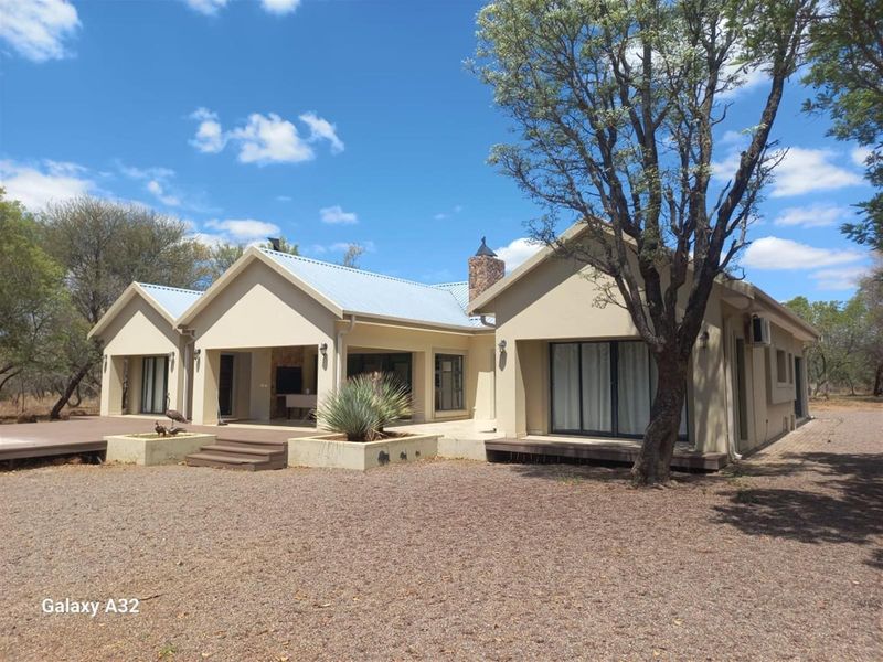 Rear view of a modern house featuring a covered patio, timber decking, and a stone chimney, set within a natural bushveld landscape.