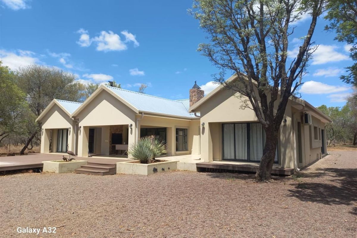 Rear view of a modern house featuring a covered patio, timber decking, and a stone chimney, set within a natural bushveld landscape.