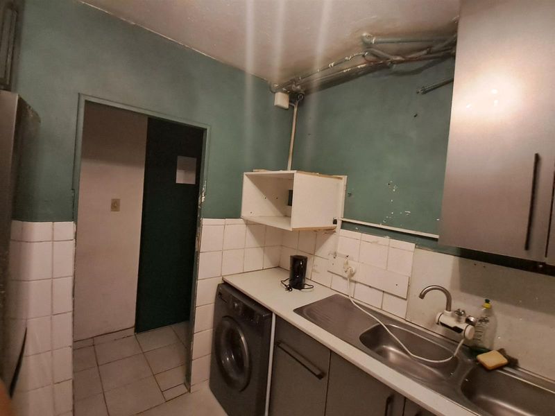 A dated kitchen area featuring a stainless steel sink, tiled backsplash, and exposed plumbing, requiring significant renovation and cosmetic updates.