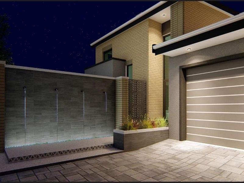 Modern exterior front view featuring a contemporary garage, stone-tiled water feature, and integrated architectural lighting at night.