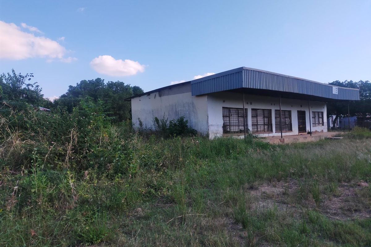 A single-story commercial building featuring a metal roof and covered veranda, set on a large plot with significant overgrown vegetation.