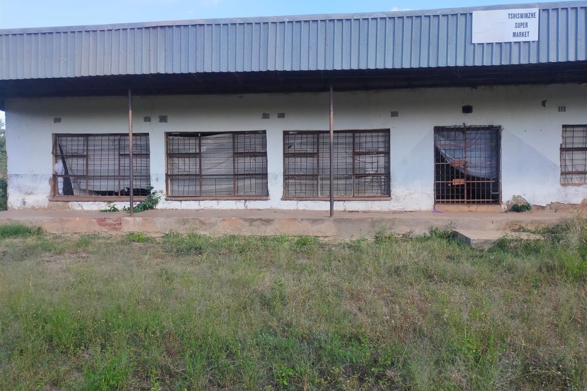 Front elevation of a commercial supermarket building featuring a corrugated metal roof, security-barred windows, and a concrete veranda.