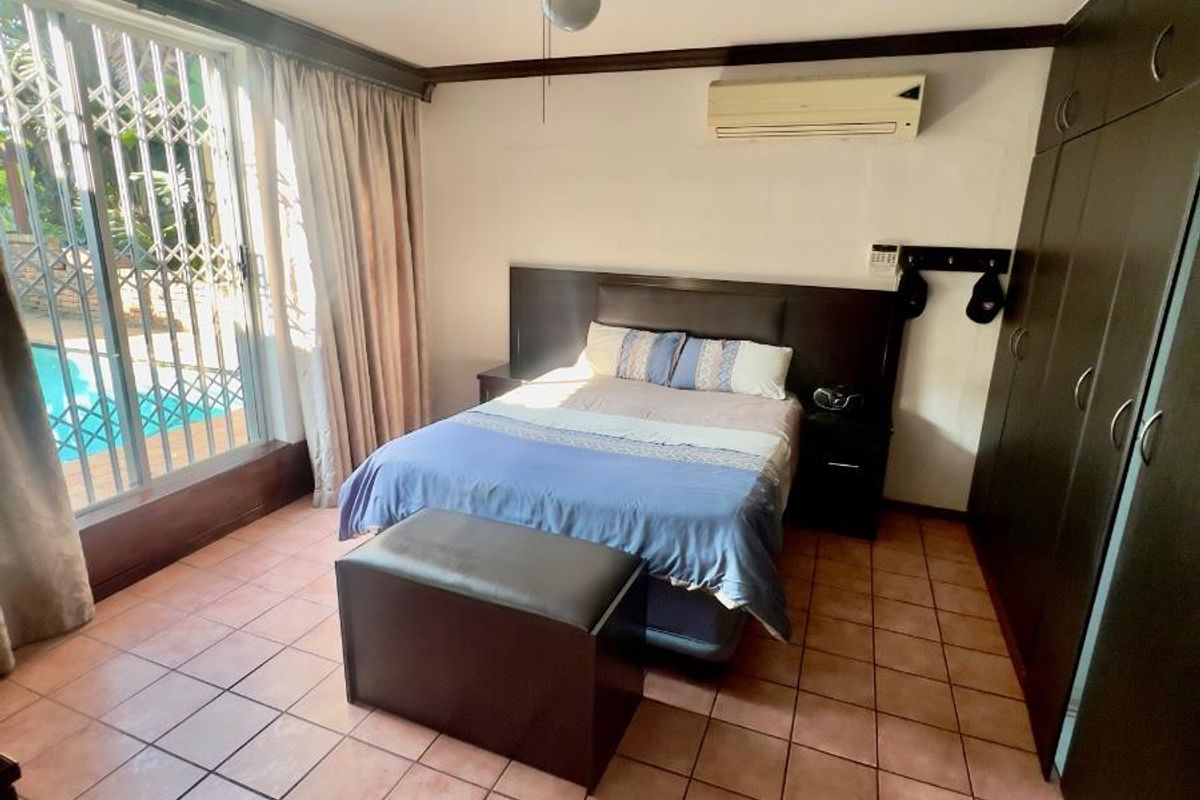 Spacious bedroom featuring built-in dark wood cabinetry, ceiling fan, wall-mounted air conditioning, and sliding glass doors with security gate leading to the pool area.