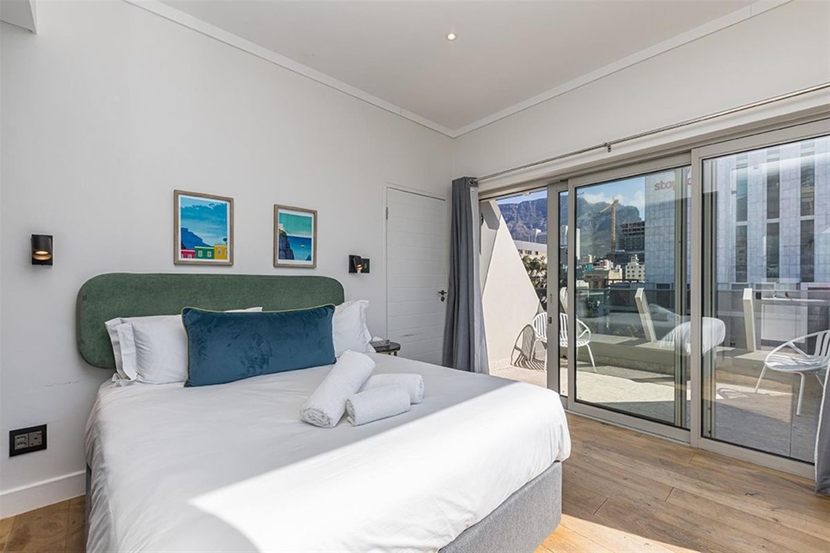 A bright bedroom featuring wood-look flooring, recessed lighting, and large sliding glass doors that open onto a private balcony with city and mountain views.