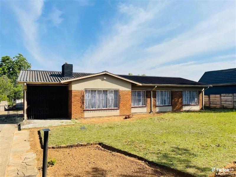 Front view of a single-story brick house featuring a tiled roof, chimney, and attached carport with security gate, set on a large grassed lot.