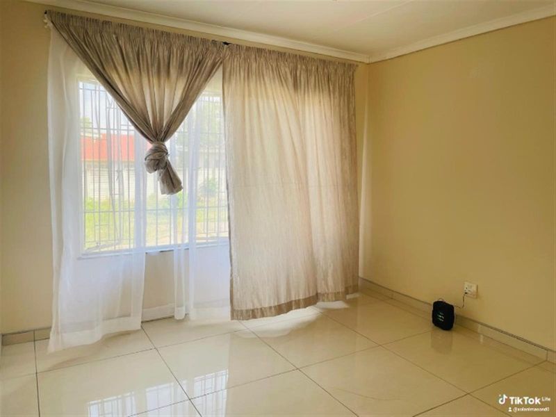 A bright, empty living area featuring neutral tiled flooring and a window fitted with security bars.