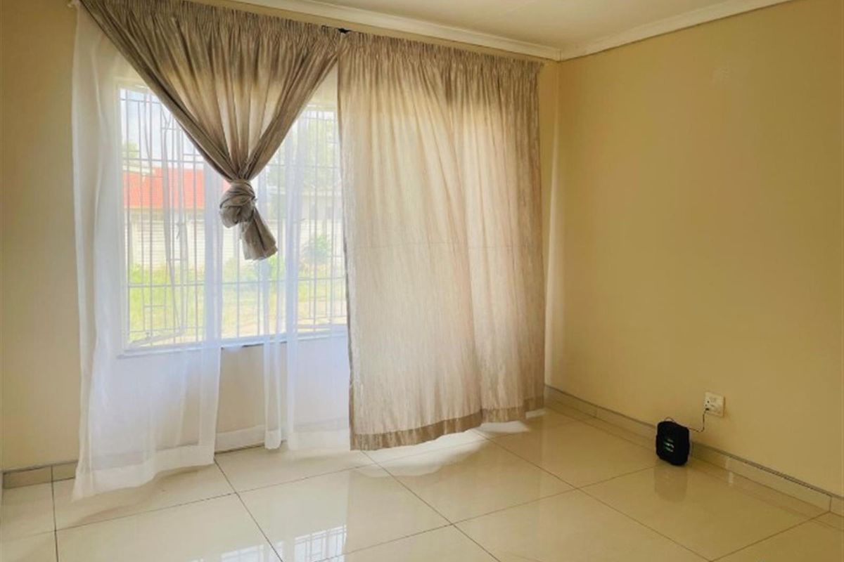 A bright, empty living area featuring neutral tiled flooring and a window fitted with security bars.