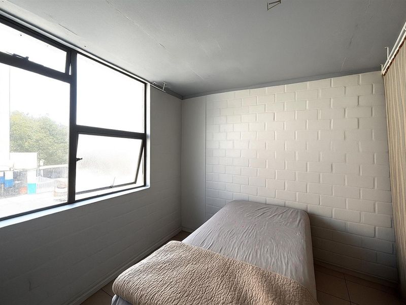A simple bedroom featuring a large window, painted brick walls, and tiled flooring, offering a blank canvas for potential interior updates.