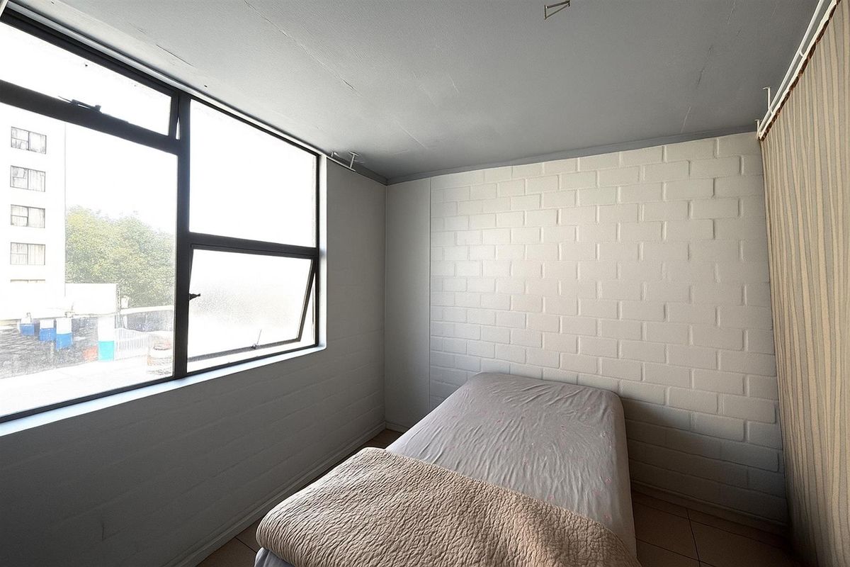 A simple bedroom featuring a large window, painted brick walls, and tiled flooring, offering a blank canvas for potential interior updates.
