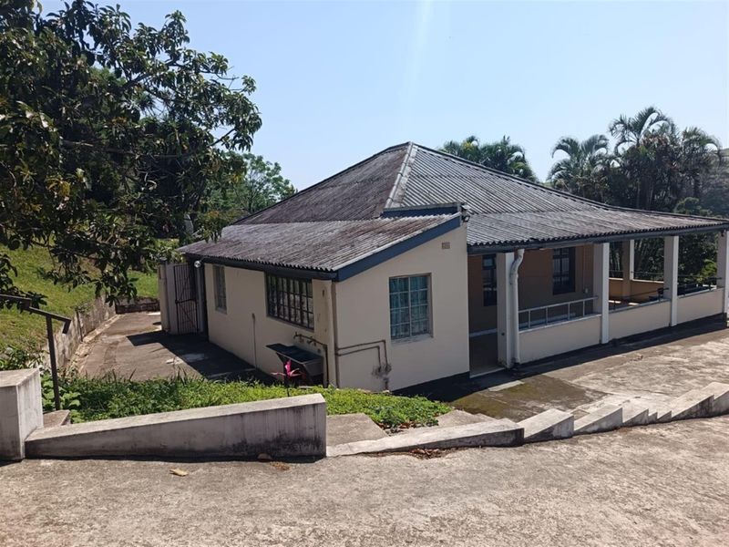 Rear view of the house featuring a covered patio, corrugated metal roof, and concrete driveway with surrounding garden area.