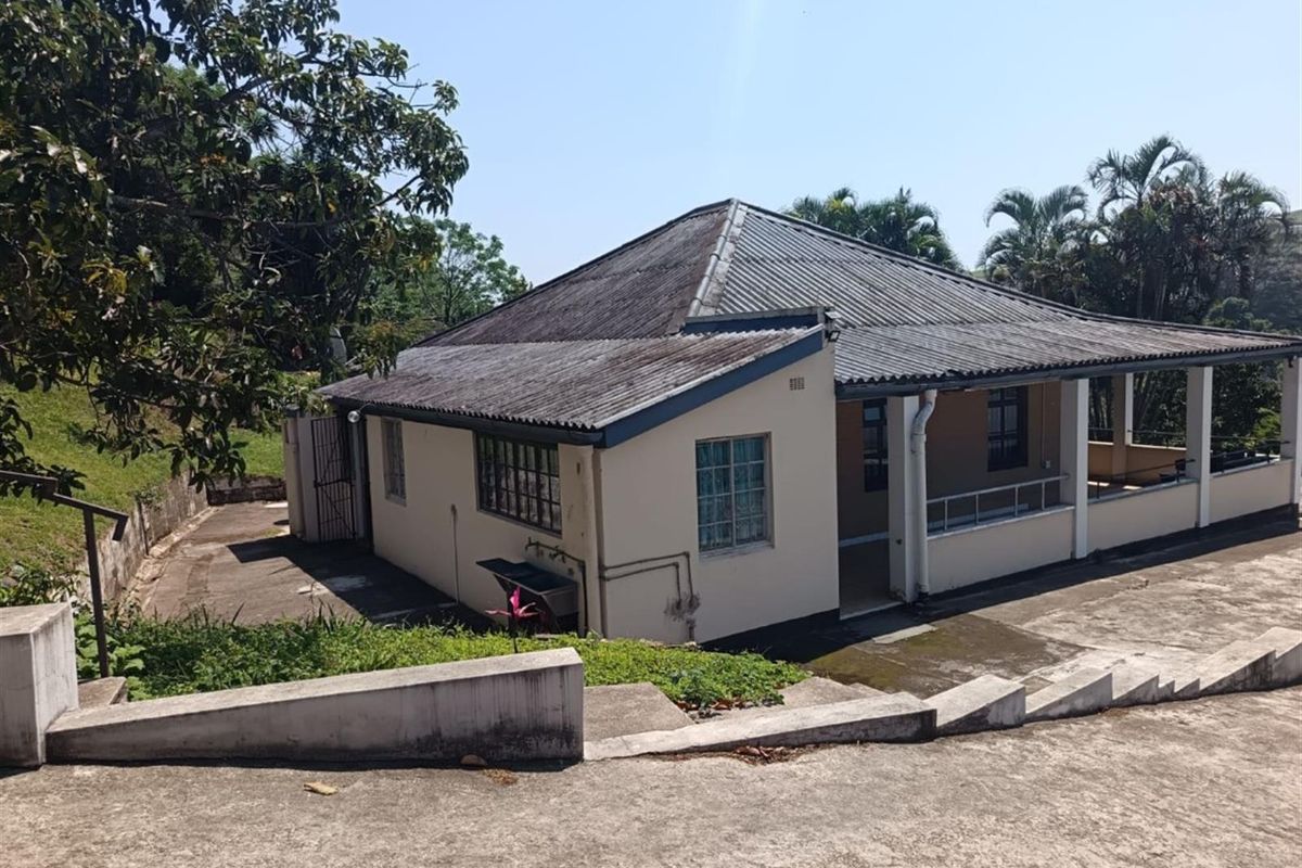 Rear view of the house featuring a covered patio, corrugated metal roof, and concrete driveway with surrounding garden area.