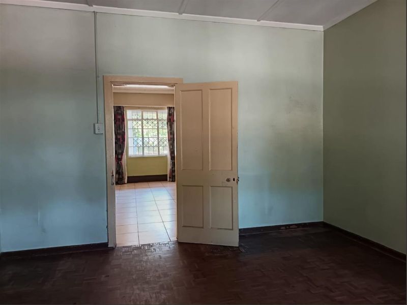 An empty bedroom featuring parquet flooring and a timber door, leading to an adjacent room with tiled flooring and security window bars.