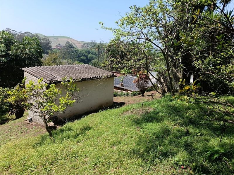 A view of a small, weathered outbuilding structure situated on a sloped, grassy garden area with surrounding trees and distant hillside views.