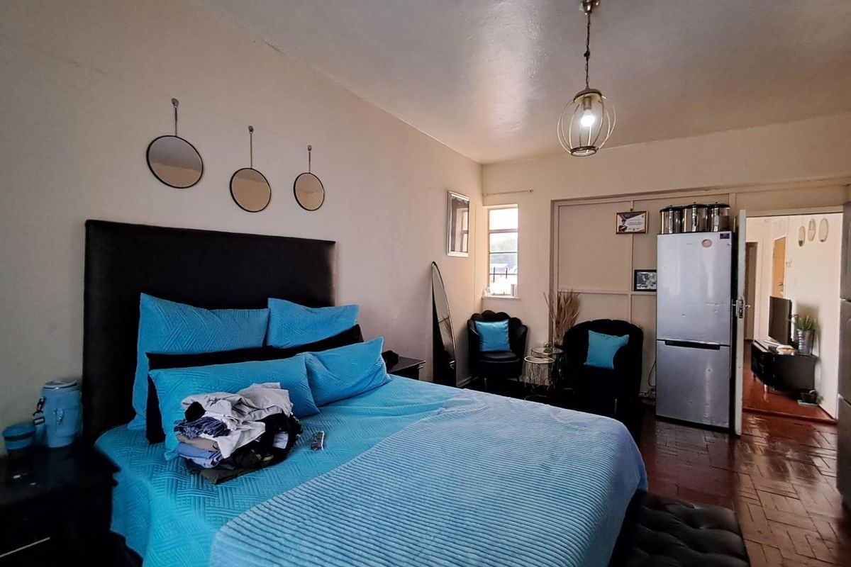 A spacious bedroom featuring wooden flooring, a pendant light fixture, and a window providing natural light, with an adjacent doorway leading to another room.