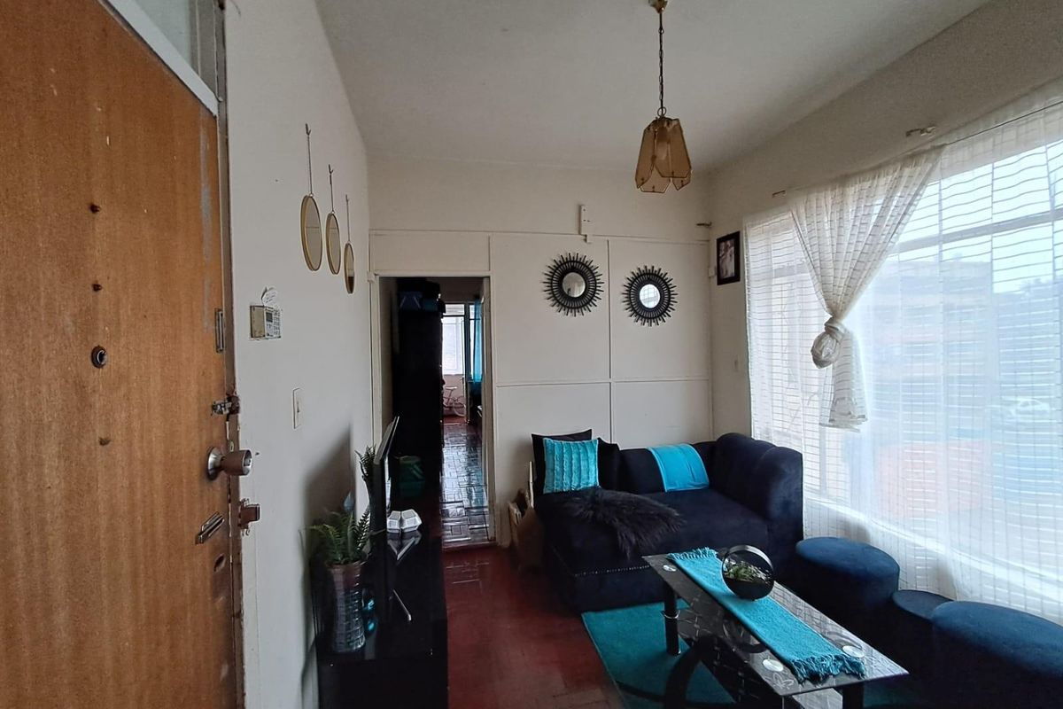 A compact living room featuring original timber flooring, large windows allowing natural light, and direct access to the hallway.