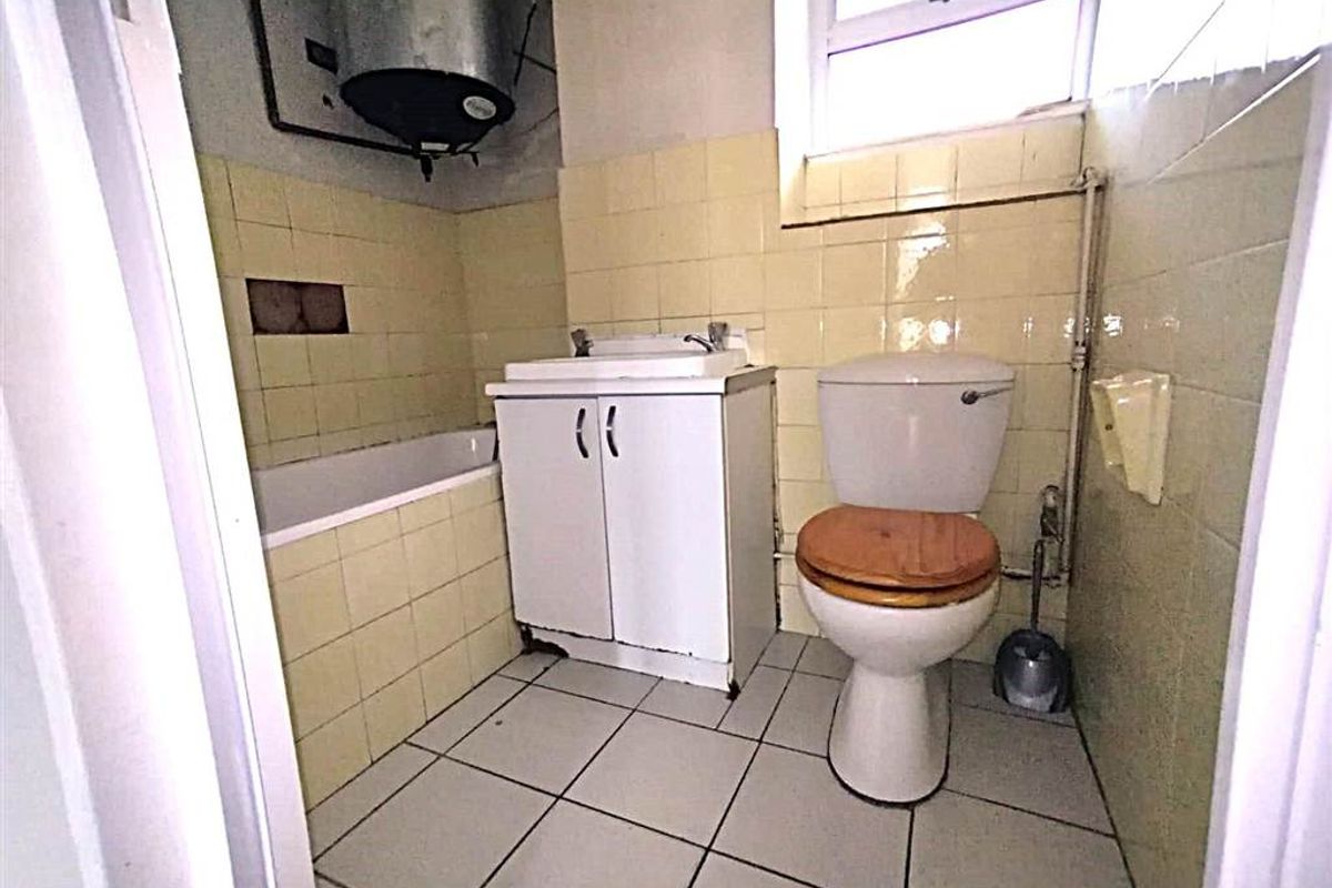 A compact, dated bathroom featuring a standard toilet, vanity basin, and bathtub with exposed water heater and original wall tiling.