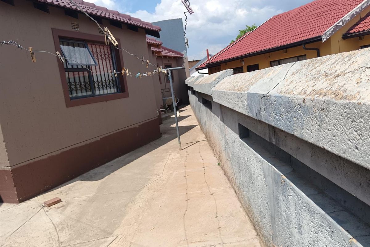 A narrow side passage between the house and a concrete boundary wall, featuring paved flooring and security bars on the window.