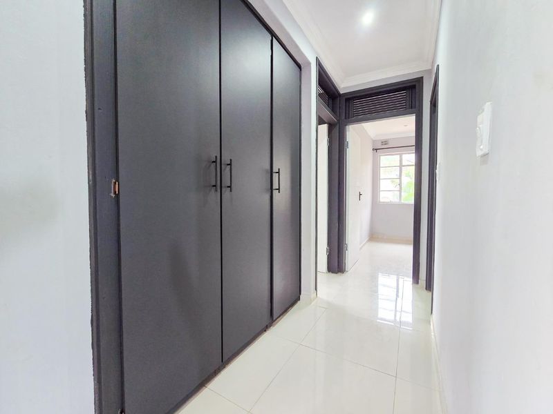 A bright, clean hallway featuring modern dark-toned built-in cupboards, white tiled flooring, and neutral walls leading to adjacent rooms.
