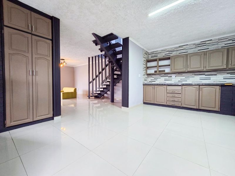 Open plan living area featuring tiled flooring, built-in kitchen cabinetry with a tiled backsplash, and a central staircase leading to the upper level.