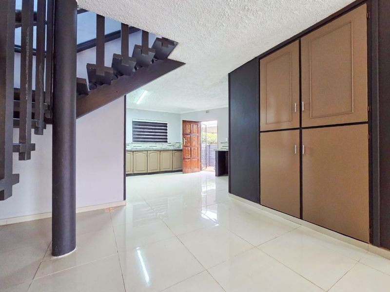 Open plan living area featuring tiled flooring, a prominent staircase, and built-in storage cupboards, leading through to a functional kitchen space.