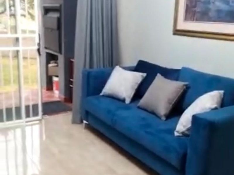 A bright living room featuring tiled flooring and a sliding security gate providing direct access to an outdoor patio area with a built-in braai.