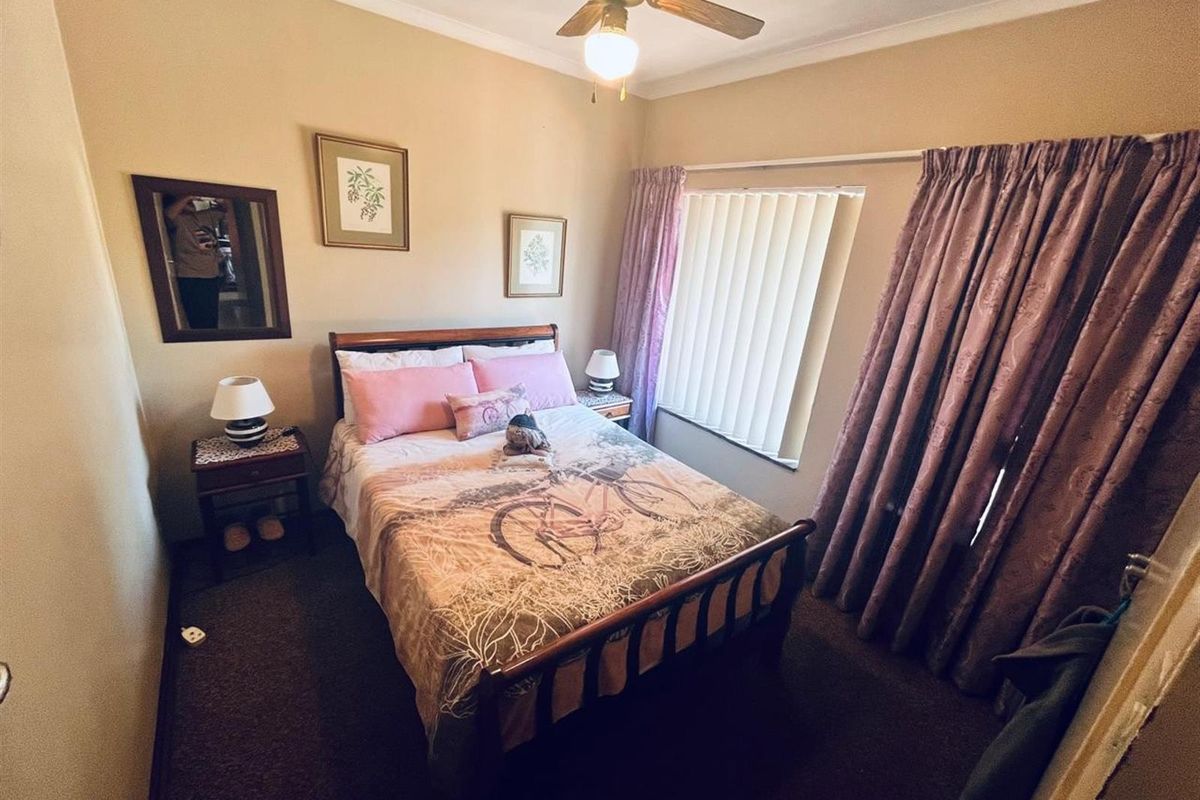 A cozy bedroom featuring a ceiling fan, carpeted flooring, and large window with vertical blinds and curtains.