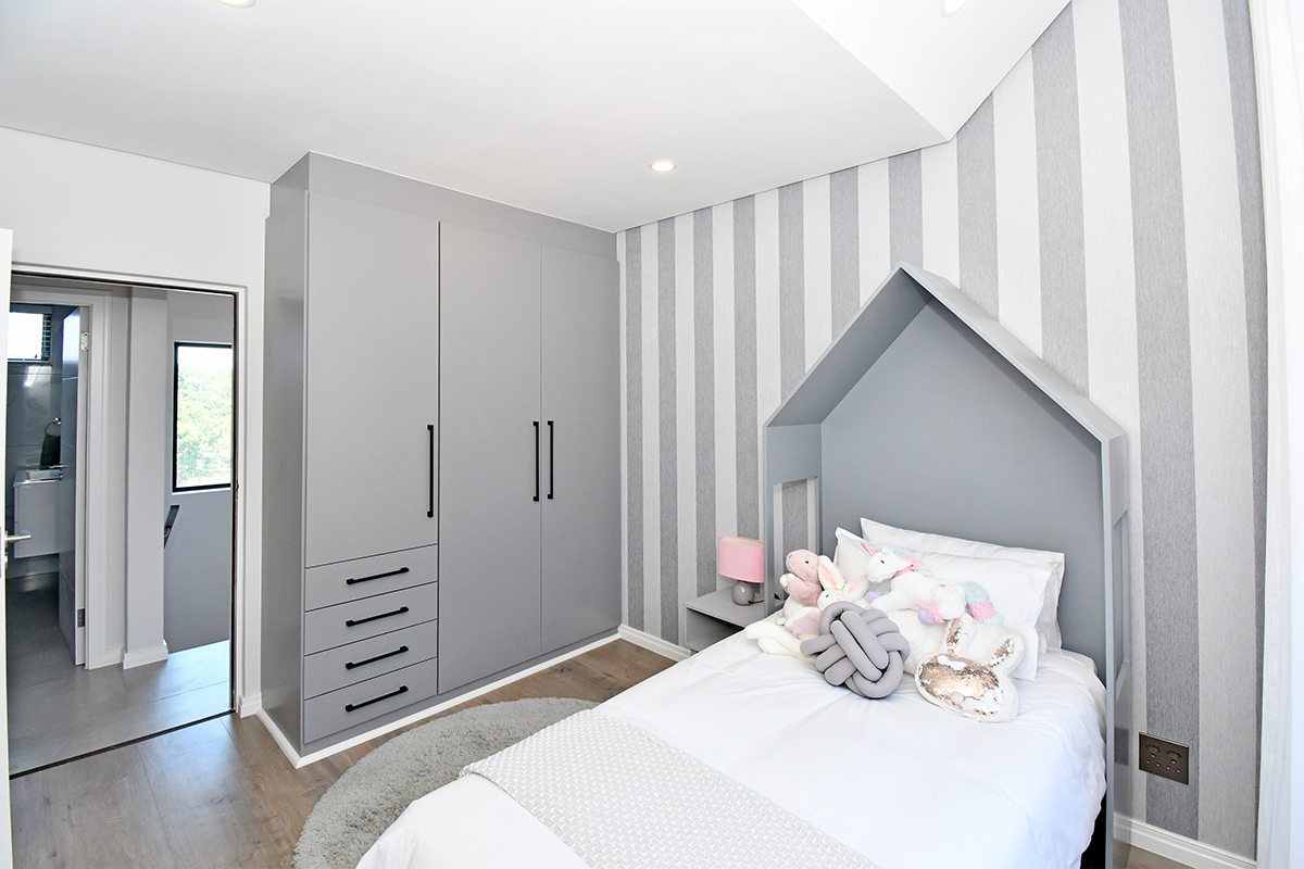 A modern bedroom featuring built-in grey cabinetry, striped feature wallpaper, recessed lighting, and durable laminate flooring, with an adjacent view into a hallway.