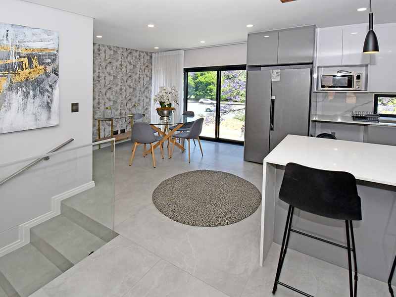 Modern open-plan kitchen and dining area featuring sleek cabinetry, a stone island, tiled flooring, and a glass balustrade overlooking the staircase.