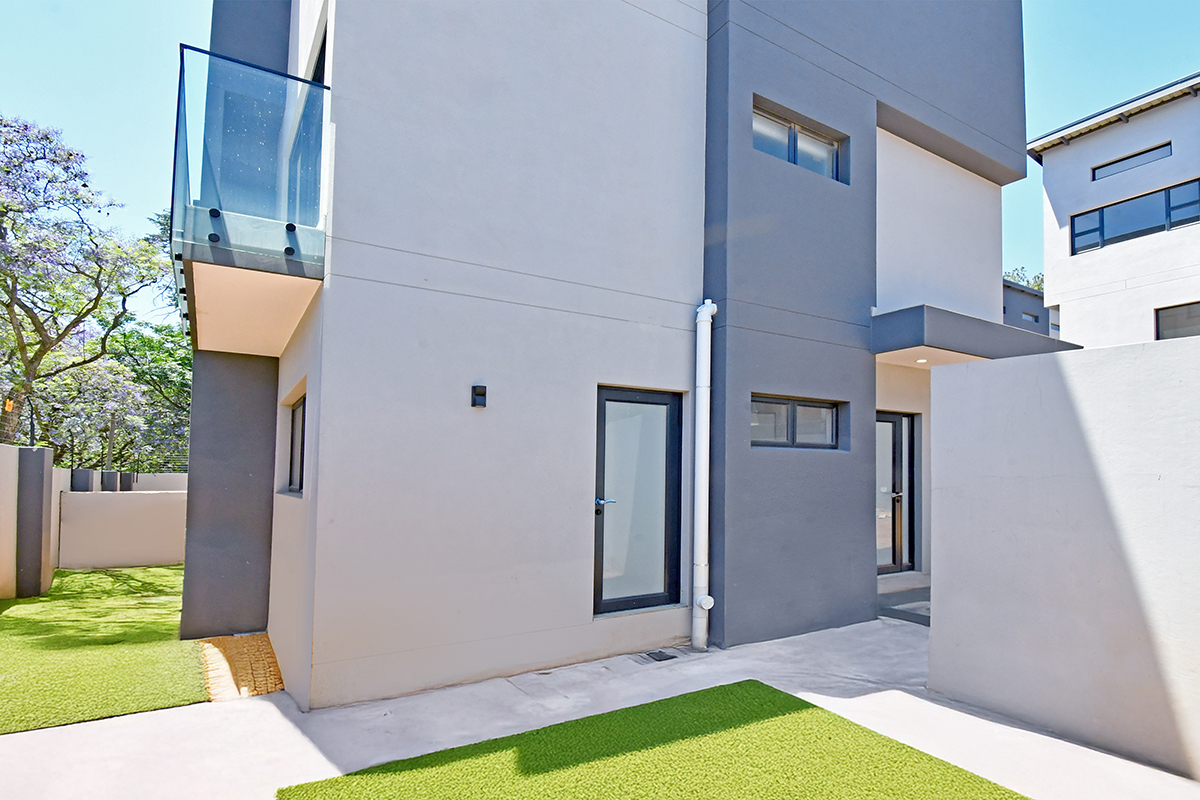 Modern rear exterior view of the property featuring a concrete patio, artificial lawn, and a glass-balustrade balcony.